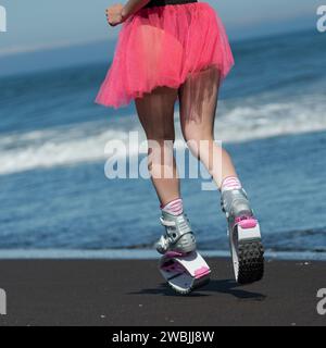 Rear view of female legs in sports Kangoo Jumps boots, swimsuit and short pink skirt running and jumping on sandy beach Stock Photo
