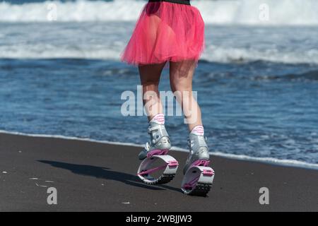 Sporty woman legs in Kangoo Jumps sports boots, black swimsuit and short skirt running and jumping on beach during Stock Photo