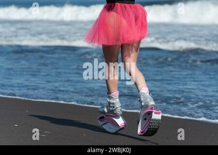 Rear view of woman legs in sports boots Kangoo Jumps, black swimsuit and short skirt running on black sandy beach Stock Photo