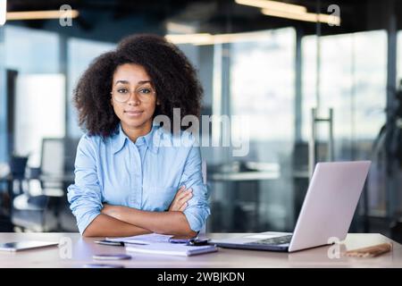 Confident and serious businesswoman looking at camera sitting at desk with crossed arms, curly haired worker using laptop at work. Stock Photo