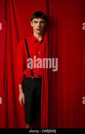 young man in vibrant shirt and shorts with suspenders hiding behind red curtain, trendy look Stock Photo