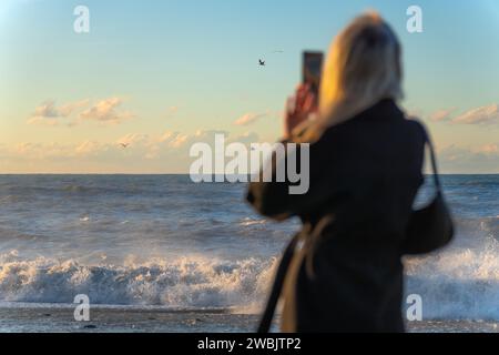 Young woman in a black coat photographs huge waves in the sea, seagulls at sunset, focus on the sea. Stormy sea and a woman with a smartphone. Stock Photo