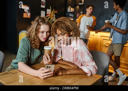 interracial girlfriends browsing internet on smartphone near coffee to go in cafe of hostel lounge Stock Photo