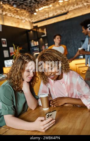 happy woman browsing social media on smartphone near african american female friend in lobby cafe Stock Photo