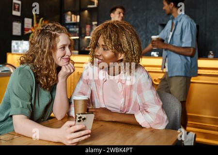 joyful interracial female friends with smartphone and coffee to go talking in lobby cafe of hostel Stock Photo