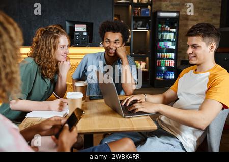 cheerful multiethnic friends talking near student networking on laptop in lounge of youth hostel Stock Photo