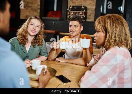 positive multiethnic friends with coffee cups talking in lounge cafe of students hostel, travelers Stock Photo