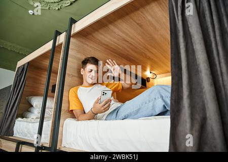 joyful student waving hand during video-call on smartphone on double-decker bed in hostel Stock Photo