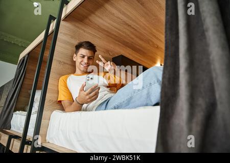 smiling student showing victory sign during video-call on smartphone on double-decker bed in hostel Stock Photo