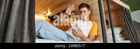 joyful guy waving hand during video-call  on double-decker bed in cozy students hostel, banner Stock Photo