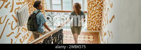 young woman and african american student with backpack walking on staircase in hostel, banner Stock Photo