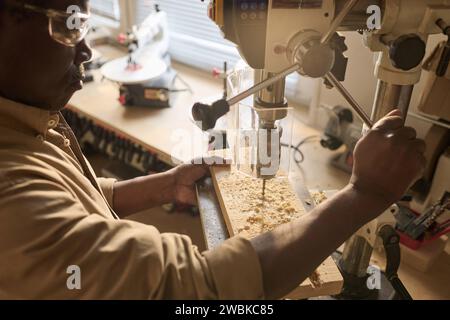 High angle closeup of Black male carpenter drilling wooden board using machine tool in workshop Stock Photo
