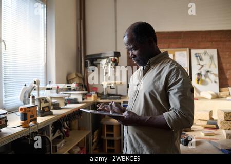 Waist up portrait of Black senior man holding digital tablet in workshop and tapping touch screen Stock Photo