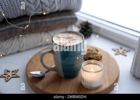 Winter windowsill still life. Blue ceramic cup of hot coffee on window sill. Christmas decorations on the background. Cozy home picture. Warm woolen k Stock Photo