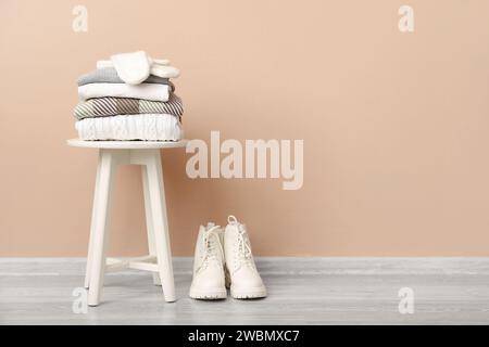 Stack of folded sweaters with gloves on stool and boots near beige wall Stock Photo