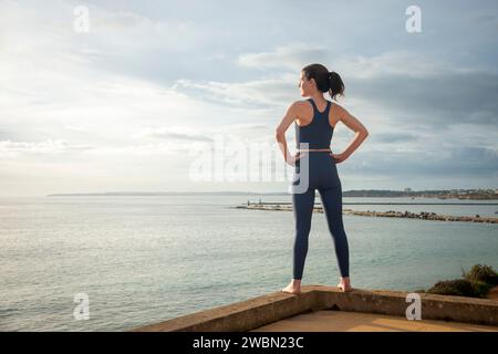 Rear view of a sporty woman facing ocean with hands on hips, relaxing after exercise. Stock Photo