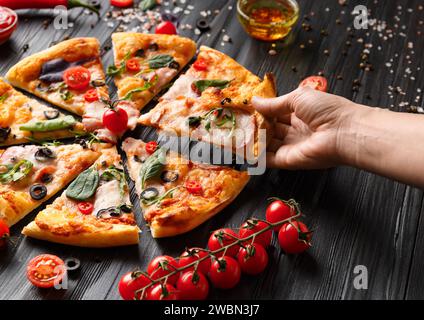 Hands take slices of cut pizza on a dark wooden background top view. Pizza with salami, jerky, sauces, spices, fresh cherry tomatoes on a black Stock Photo