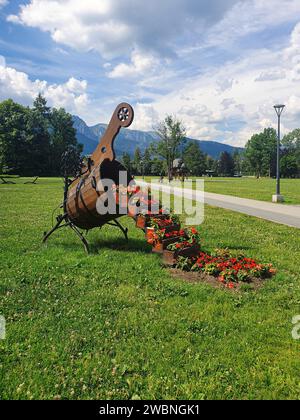 In Zakopane, a large wooden vase on green grass, a staircase full of small vases with colorful flowers, a post, trees, mountains, blue sky and clouds. Stock Photo