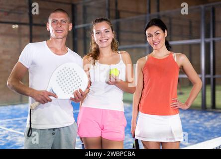 Smiling man and women with rackets and balls posing on indoor padel court Stock Photo