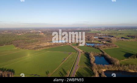 This aerial image captures the expansive beauty of the countryside, characterized by lush green fields that stretch into the distance, suggesting fertile agricultural land. A series of small lakes or ponds adds a touch of blue to the verdant landscape, enhancing its picturesque quality. A country road meanders through the fields, offering connectivity through this bucolic setting. In the backdrop, a hint of urban development on the horizon provides a subtle contrast to the rural foreground. The clear skies and the soft light of a day nearing its end cast long shadows and enrich the colors of t Stock Photo