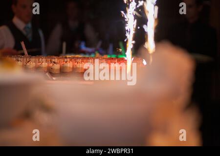 This photograph captures a festive moment at a celebration, with a focus on a dessert display featuring an array of delicacies, each topped with a lit sparkler. The foreground is softly blurred, emphasizing the sparkling lights and their bright, energetic trails. In the background, guests are gathered, their silhouettes and the ambient lighting contributing to the convivial atmosphere. The warm and inviting glow of the sparklers suggests a cheerful occasion, possibly a wedding or a grand party, where dessert is served with a side of spectacle. Sparkling Celebration: A Festive Dessert Display.  Stock Photo