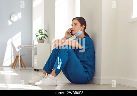 Young nurse or doctor having a break, sitting on the floor and talking on her mobile phone Stock Photo