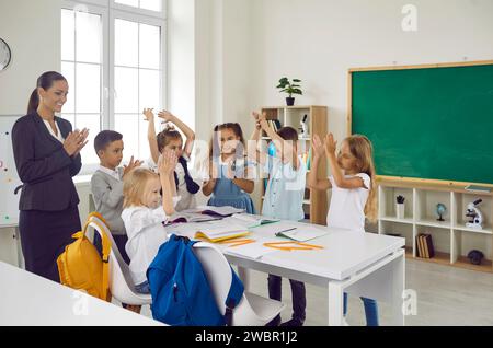 Happy children and woman teacher applaud stand around school desk with notebooks and pencils Stock Photo