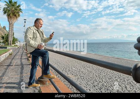 Smiling middle-aged bearded man drinking coffee and using smartphone on seaside in sunny winter or spring day. Stock Photo