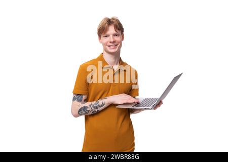 a young successful red-haired guy dressed in an orange t-shirt with a tattoo on his arm works as a programmer and holds a laptop in his hand Stock Photo