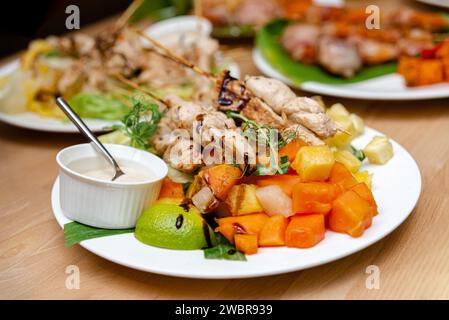 Grilled chicken skewers garnished with fresh herbs, served with a colorful tropical fruit salad and a side of creamy dipping sauce. Stock Photo