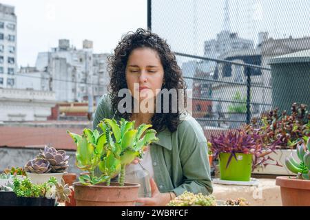 young latin woman in curlers, standing at work maintaining plants on terrace, using water sprayer to hydrate them, home work concept, copy space. Stock Photo