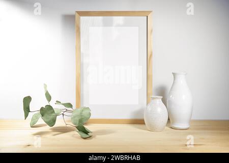 Mockup of an empty picture frame, two white ceramic vases and an eucalyptus branch on a wooden table against a light gray wall, minimal Scandinavian s Stock Photo