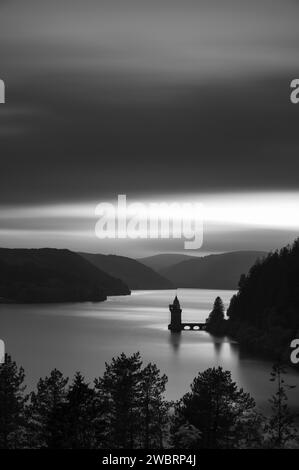 Long exposure, black and white, of Lake Vyrnwy, in Mid Wales. There is a pumping station jutting out into the lake, whcih is surrounded by lush, green Stock Photo