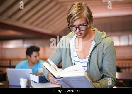 Reading book, knowledge or student in library at university, college or school campus for education. Glasses, nerd or male person serious about Stock Photo