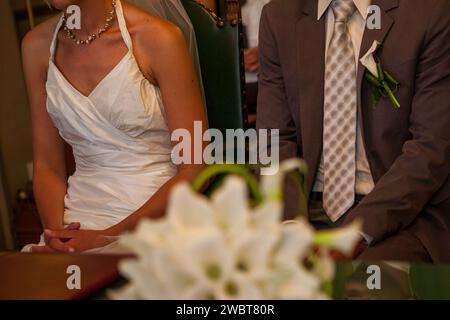 This photograph captures a nuanced moment at a wedding, focusing on the mid-section of the bride and groom as they sit side by side. The bride's elegant dress and the groom's formal attire, along with a glimpse of their posture, suggest the dignified poise of the occasion. A wedding flower arrangement softly blurs in the foreground, framing the couple's folded hands, which are gently resting on the bride's lap. This detail subtly conveys the connection and mutual support that characterize their bond. The intimacy of the setting is heightened by the soft lighting and close framing, which leave  Stock Photo