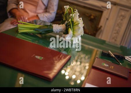 This photograph captures the poetic stillness of a bridal bouquet resting on the signing registry, a moment frozen in the midst of matrimonial proceedings. The bouquet, composed of white lilies and adorned with a delicate lace, is positioned prominently in the foreground, symbolizing the purity and beauty of the wedding occasion. In the soft-focus background, we see a bride in a pale suit, her posture reflecting the gravity and joy of the event. The green table and the reflective surface add to the elegance of the setting, highlighting the significance of the documents that will seal the union Stock Photo