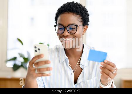 Happy young attractive multiracial woman shopping online via smartphone, using credit card at office space, african-american charismatic businesswoman with teeth gap making money transfer, e-banking Stock Photo