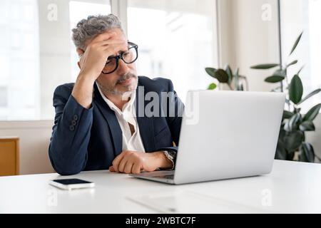 Thoughtful male office employee using laptop indoors, looking at laptop screen with puzzled face expression, businessman solving tasks, having problems with finance Stock Photo