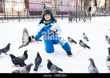 boy in a fur hat and warm overalls has fun surrounded by pigeons while walking in the city on a winter day. Many wild gray pigeons fly to the hands of Stock Photo