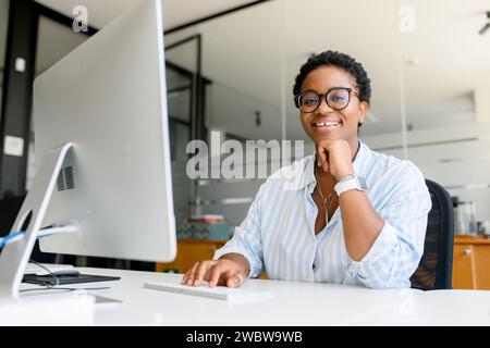 Positive African American female employee, freelancer or programmer in glasses sitting at desk with PC and typing, writing article, woman smiling and looking at camera, enjoying work at this company Stock Photo