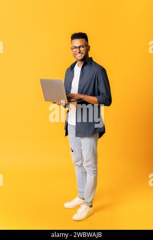 Full length portrait of nice smiling indian male entrepreneur using laptop standing isolated on yellow, happy determined male student looking at camera, holding computer, arab man typing, vertical Stock Photo
