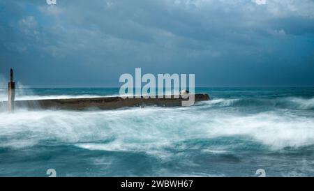 The image captures the powerful force of waves crashing against the rocks along a pier Stock Photo