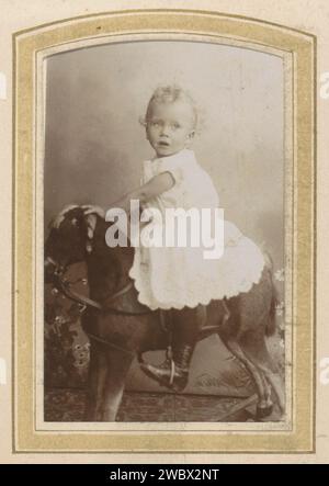 Portrait of a toddler in a dress on a rocking horse, Cornelis Johannes Lodewicus Vermeulen, c. 1890 - c. 1915 Photograph. visit card Part of photo album of a Dutch family with 168 cartes-de-visiting and cabinet photos. Amsterdam cardboard. paper. photographic support  historical persons. studio requisites  photographer. child. clothes covering the entire body. gear for legs and feet. (playing with) toys Amsterdam Stock Photo