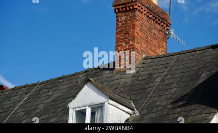 Old tiled rooftop with slates and a tall red brick chimney stack set against a blue sky with copy space Stock Photo