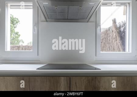 Kitchen with black ceramic hob on the wall without backsplash, stainless steel extractor hood, two windows overlooking a patio and furniture with wood Stock Photo