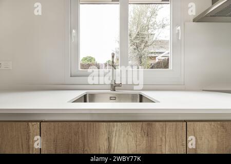 A small square sink in a kitchen with oak cabinets and white stone worktop beneath a white aluminum double window Stock Photo