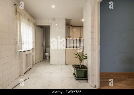 Empty kitchen with old white tiles, a window with curtains, a small bathroom in the back and an empty room on the right Stock Photo