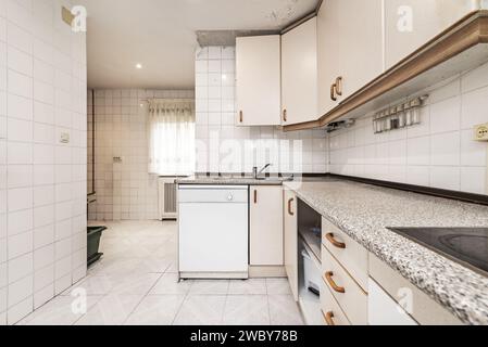 kitchen with white tiles with L-shaped furniture, a window with curtains and similar granite countertop Stock Photo