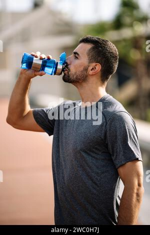 Young sports man drinking water from a bottle resting after workout in a park. Healthy fit male drinks and recovery with refreshment after exercise Stock Photo