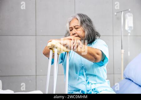 An Asian woman sitting on a bed, leaning on  crutches in a hospital Stock Photo